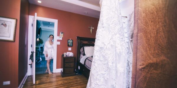 A bride-to-be stands near a wedding dress in a bedroom.
