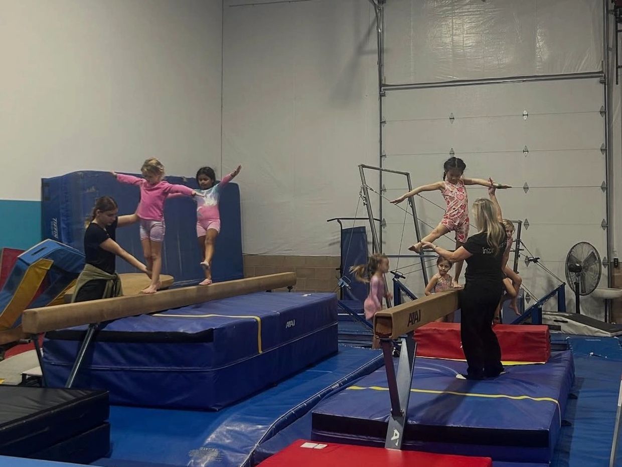 Group of children doing kicks on balance beam with coaches helping