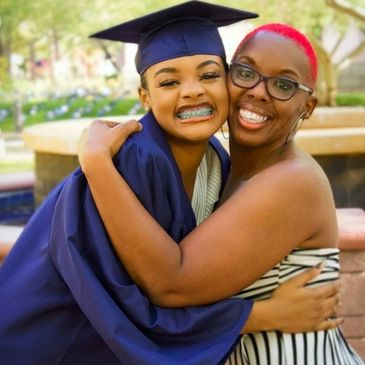 Joyful graduate in cap and gown hugging a smiling woman outdoors.