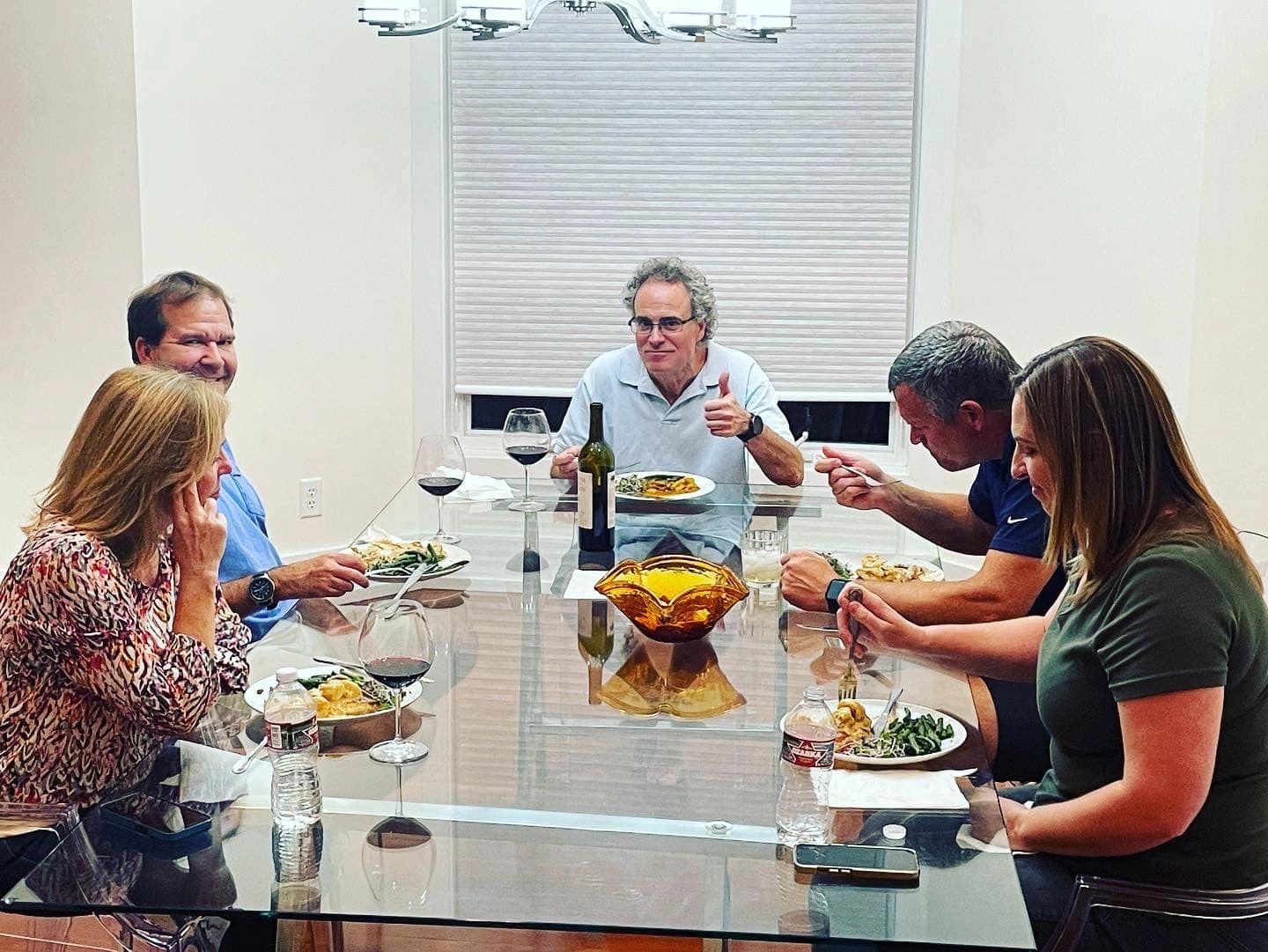 Five people enjoy dinner together around a glass table with wine and water.