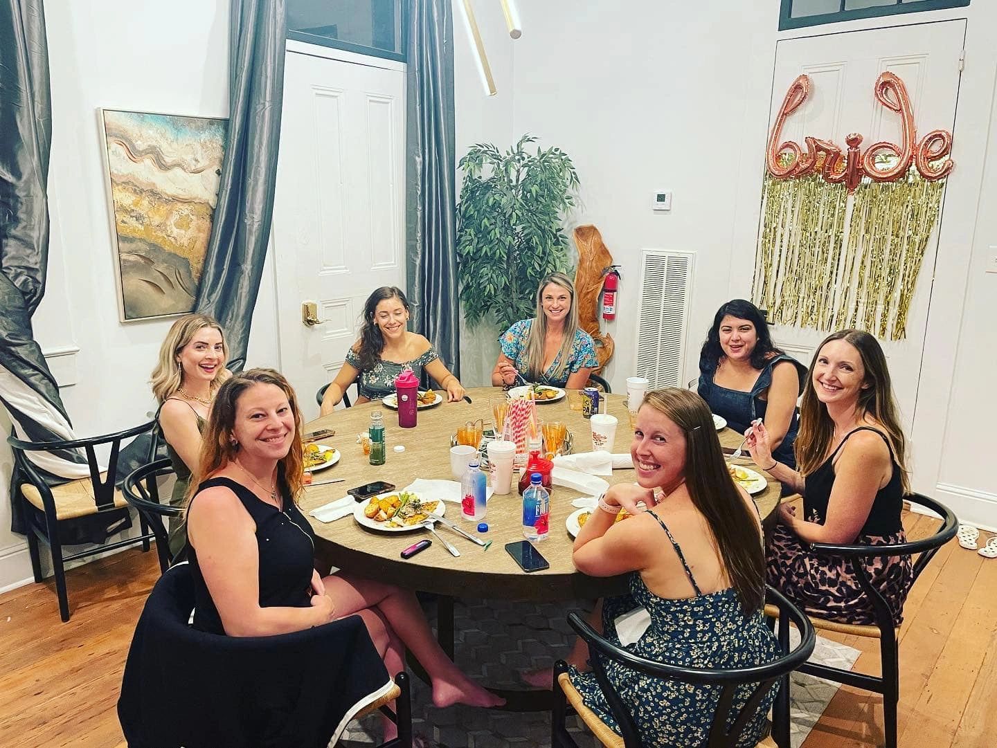 Seven women smiling around a table with food, celebrating a bride-to-be.