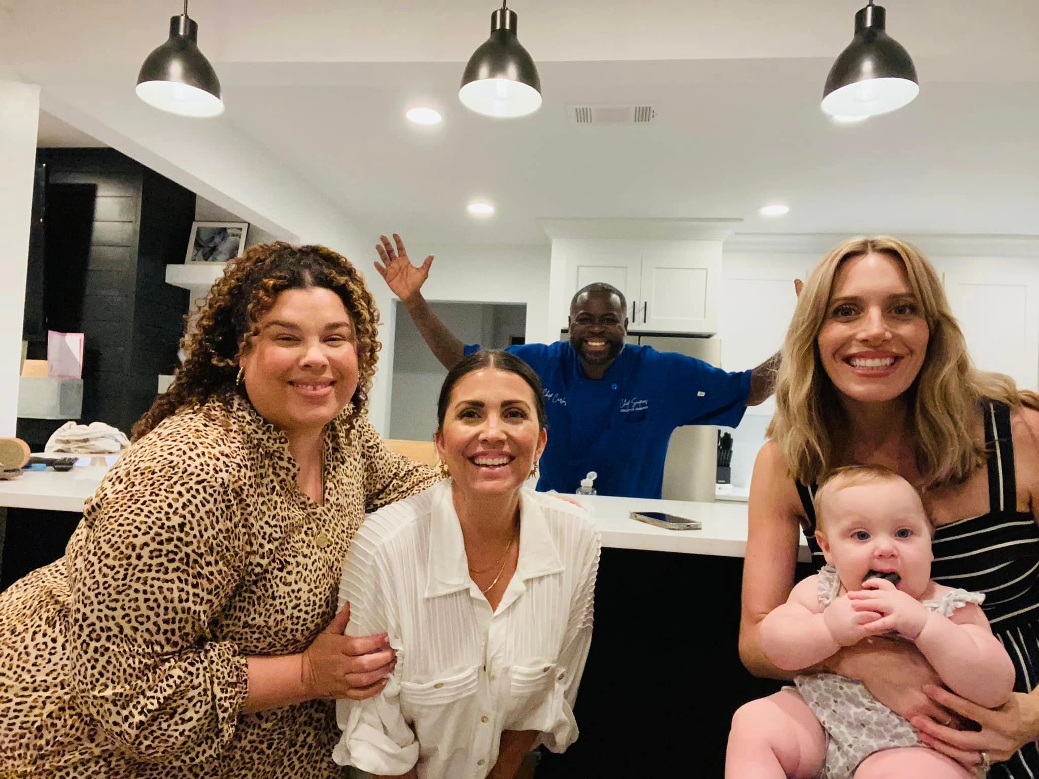 Four adults and a baby smiling together in a modern kitchen.