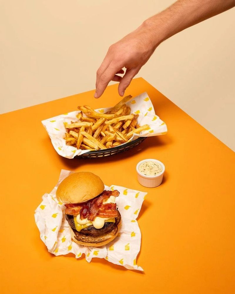 Hand reaching for fries next to a bacon cheeseburger and sauce on an orange table.