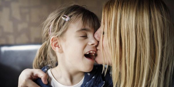 A mom gently kissing her special needs daughter on the cheek as she smiles.