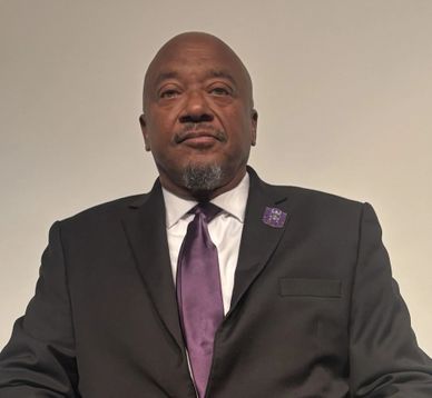 Man in a black suit with a purple tie and lapel pin.