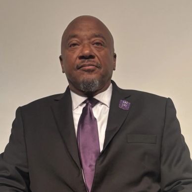 Man in a black suit with a purple tie and lapel pin.