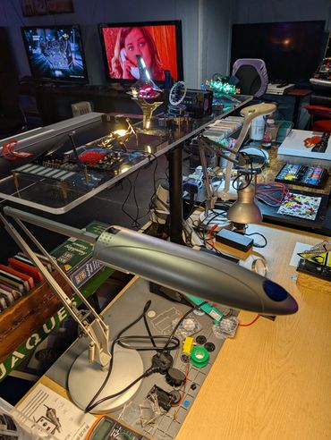 A cluttered workspace with desk lamps, electronics, and a TV playing in the background.