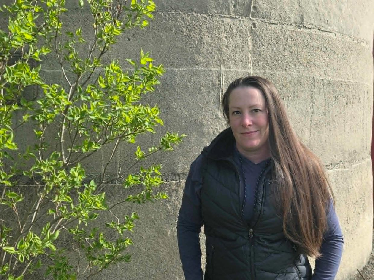 Woman with long hair standing outdoors near a tree and concrete wall.