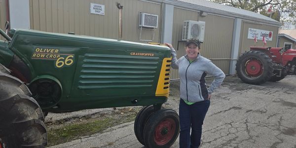 A woman stands smiling beside a vintage Oliver Row Crop 66 tractor.