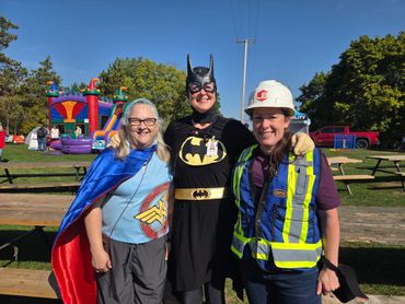 Three women dressed as Wonder Woman, Batwoman, and a construction worker pose at an outdoor event.