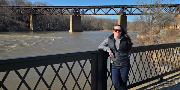 Woman standing by a river with a bridge in the background on a sunny day.