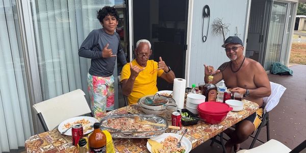 Three men at an outdoor table enjoying food and drinks, making shaka hand signs.