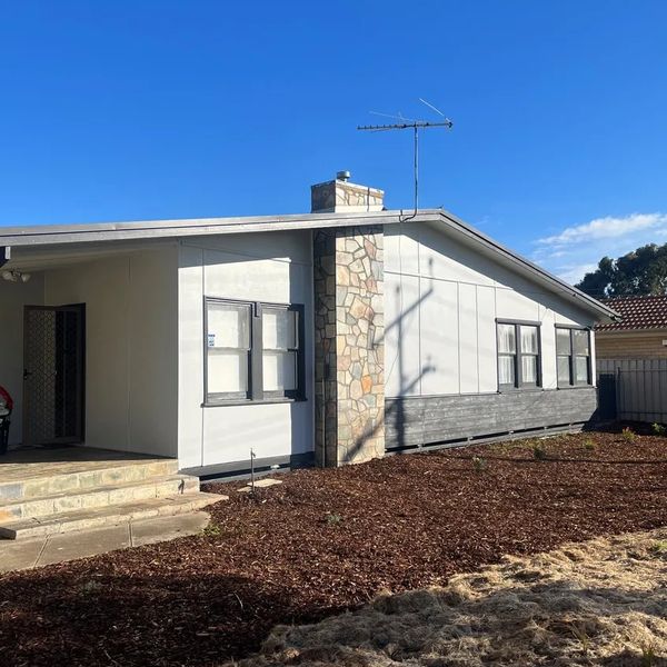 Single-story house with stone chimney and freshly mulched yard under clear blue sky.