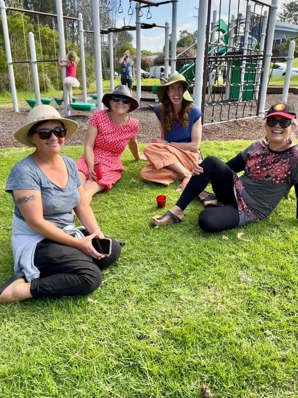 three smiling women sitting on the lawn in front of a playground