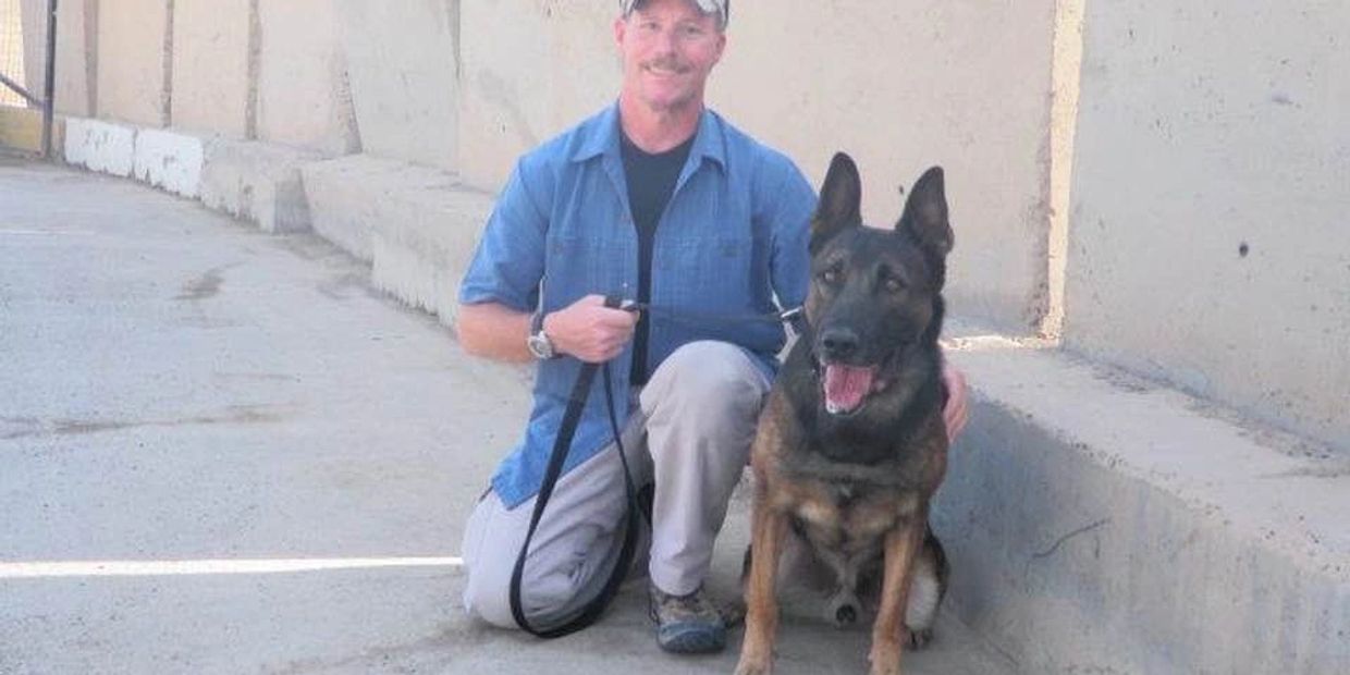 Man kneeling with a German Shepherd dog on a leash by a concrete wall.