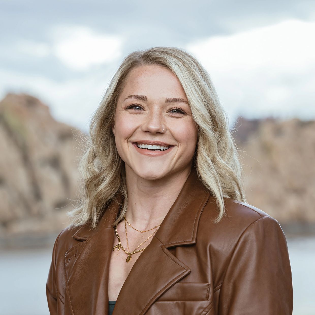 Smiling woman with blonde hair wearing a brown leather jacket outdoors.