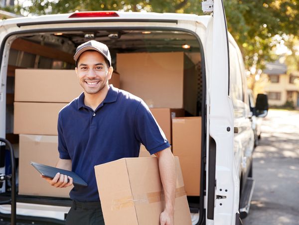A delivery person stands outside a van, holding a tablet and a cardboard box.