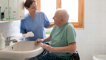 Nurse assisting elderly man in a wheelchair by the sink.
