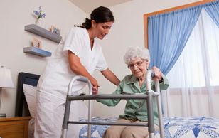 Caregiver assisting elderly woman with a walker in bedroom.