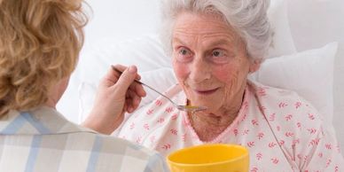 Caregiver feeding elderly woman in bed with a spoon and yellow bowl.