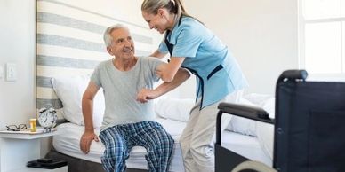 A nurse helps an elderly man sit up on a bed in a bright room.