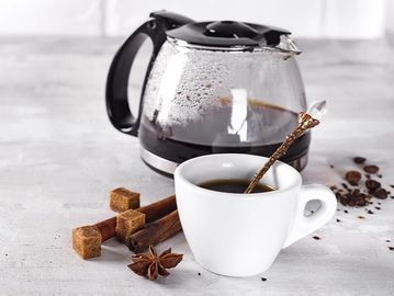 A steaming cup of coffee with a glass pot and cinnamon sticks.