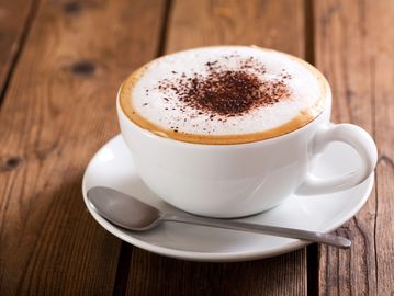 A cup of cappuccino with frothy milk and cocoa powder on top, served with a spoon.