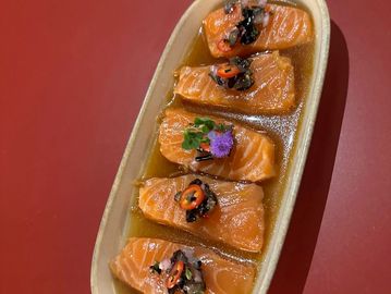 Five pieces of salmon sashimi with garnishes on a beige plate.