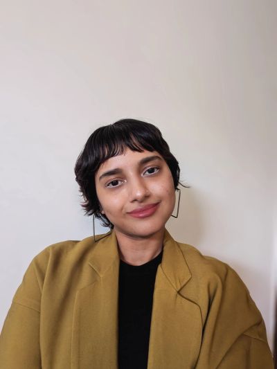 Person with short black hair wearing a mustard blazer and black top.