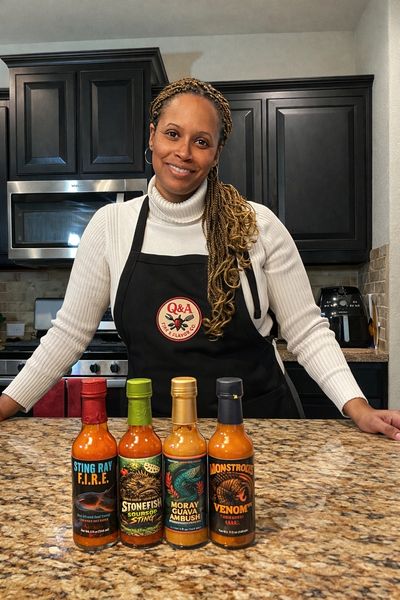 Woman in kitchen with four hot sauce bottles on counter, wearing Q&A apron.