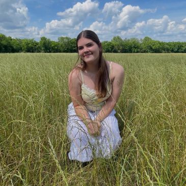 Young woman kneeling in a grassy field under a partly cloudy sky.