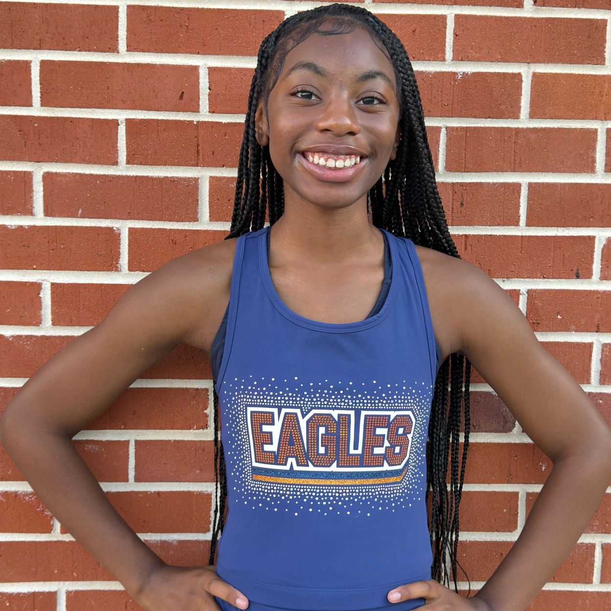 Smiling young woman in a blue "Eagles" athletic top stands confidently against a brick wall.
