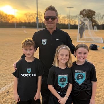A man with three children in soccer uniforms at sunset on a field.