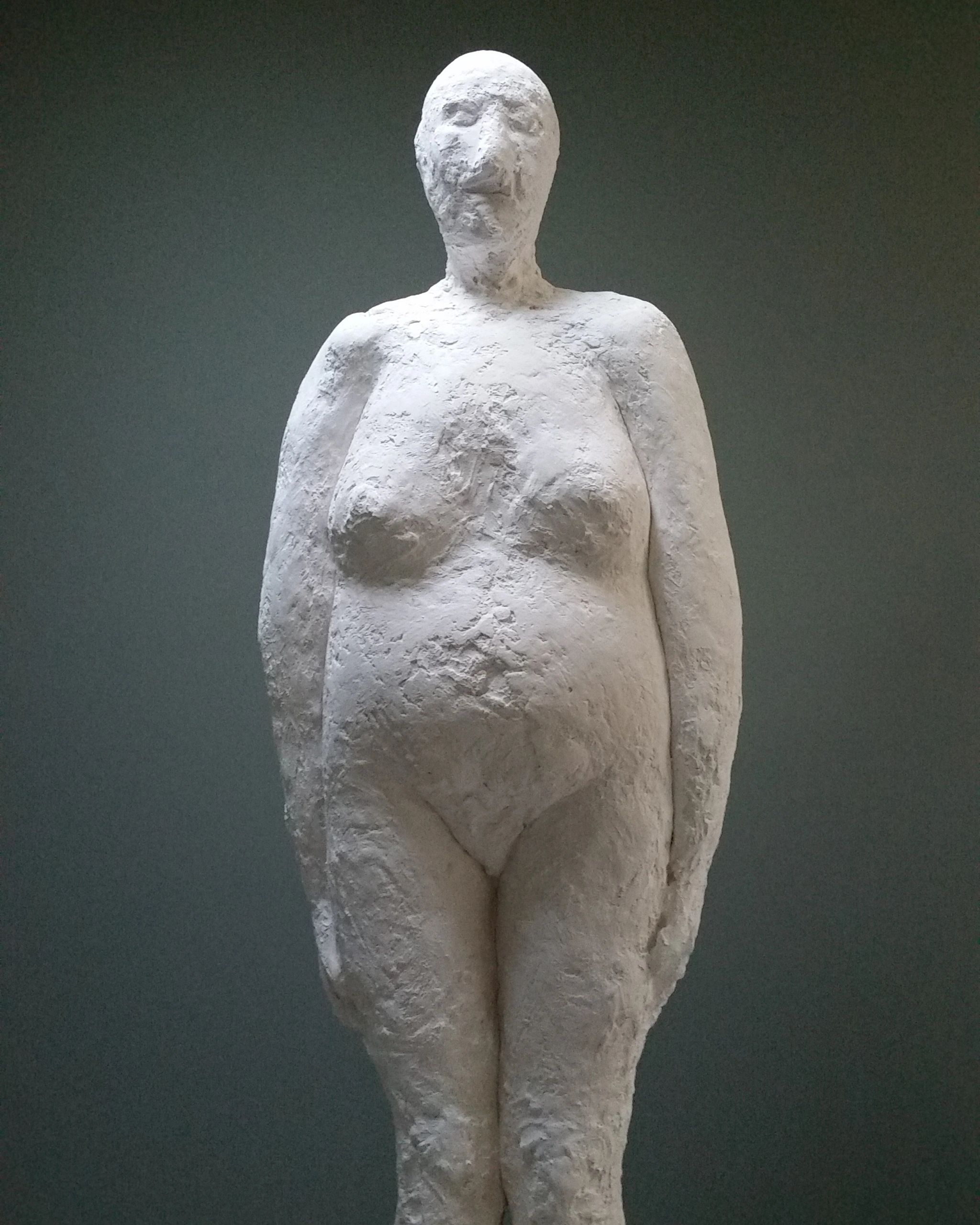 Rough-textured white sculpture of a standing human figure against a dark background.