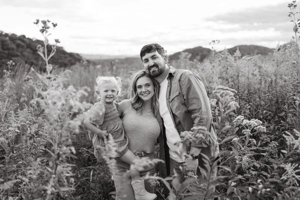 A happy family of three smiling in a field with mountains in the background.