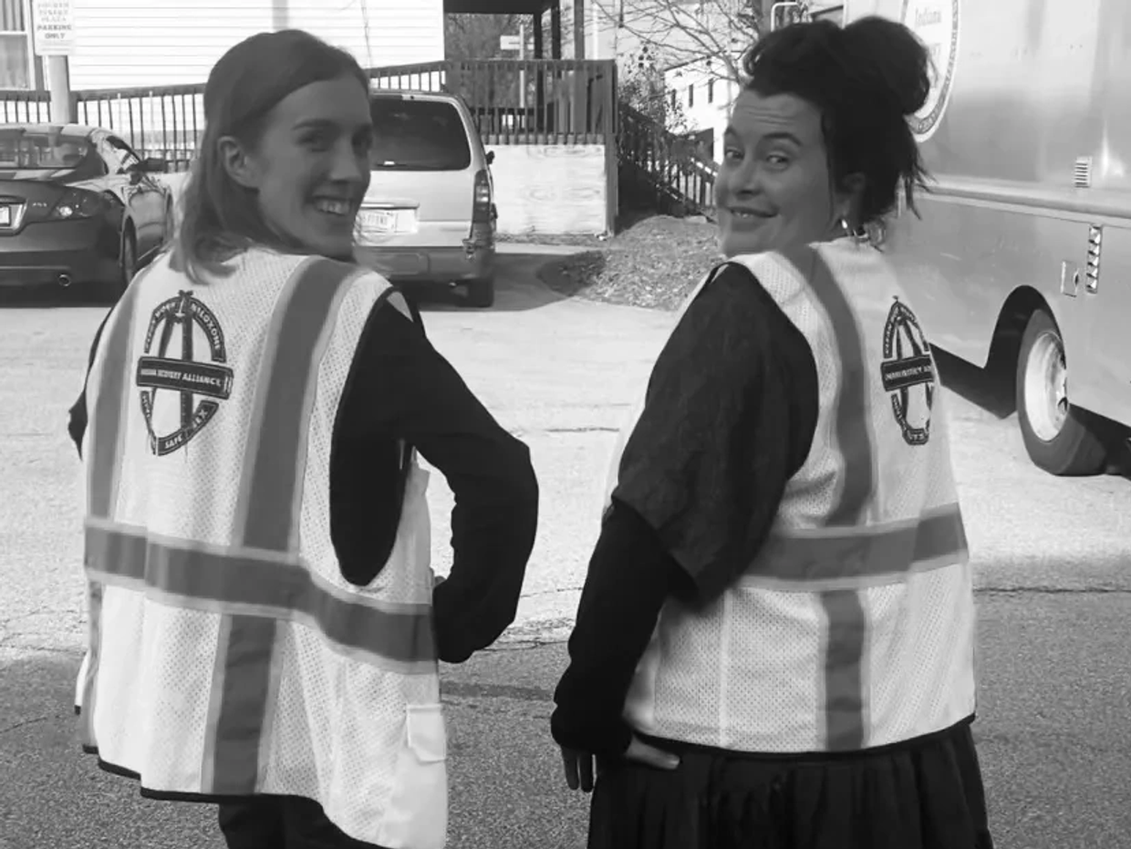 Two women in safety vests smiling and looking back outdoors.