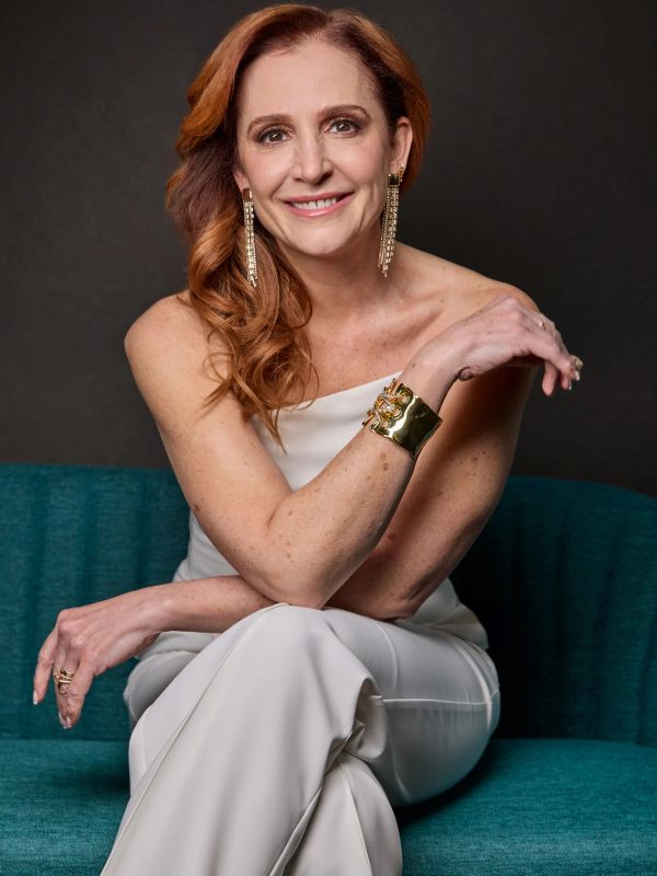 Elegant woman with red hair wearing white and gold jewelry, sitting on a teal sofa.