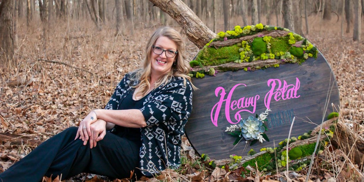 Woman sitting in forest beside a decorated wooden sign reading 'Heavy Petal'.