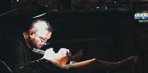A tattoo artist working on a client's leg in a dimly lit studio.