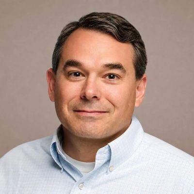Professional headshot of a middle-aged man with dark hair and a light blue shirt.