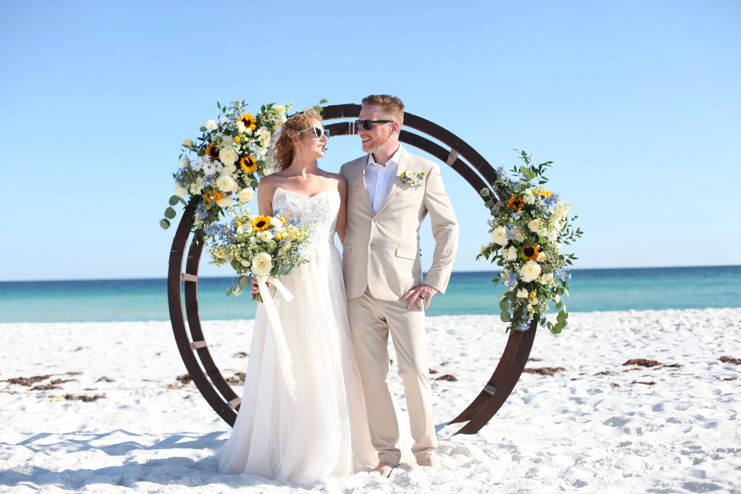 Couple in wedding attire on a beach with floral decorations.