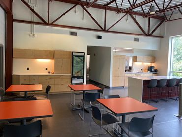 Modern office kitchen and dining area with orange tables and black chairs.
