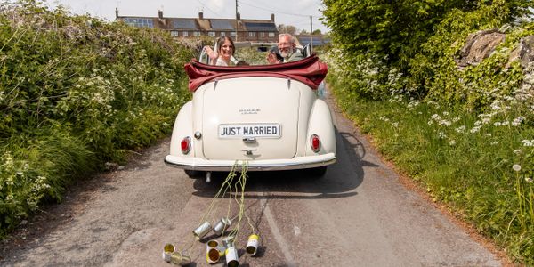 Newlyweds driving away in a vintage car with a 'Just Married' sign and tin cans tied to the back.