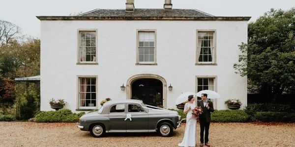 A newlywed couple stands with umbrellas in front of a vintage car and a large white house.