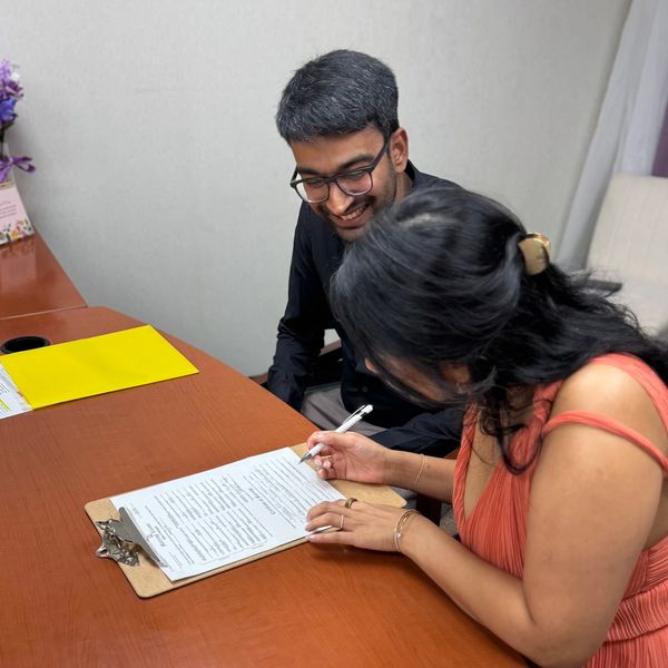 A woman signing a document while a man watches and smiles.