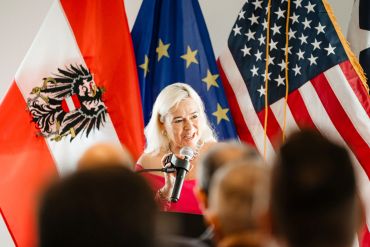 Woman speaking at a podium with Austrian, EU, and US flags.