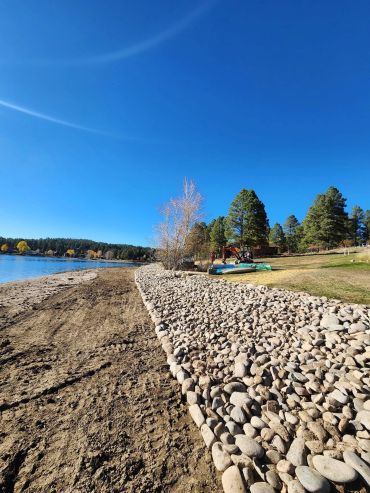 A lakeside shore with rocks, trees, and clear blue sky.