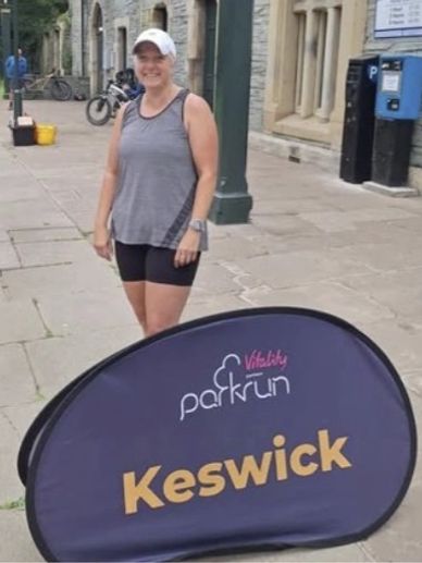 Smiling woman in sportswear near Keswick parkrun sign.