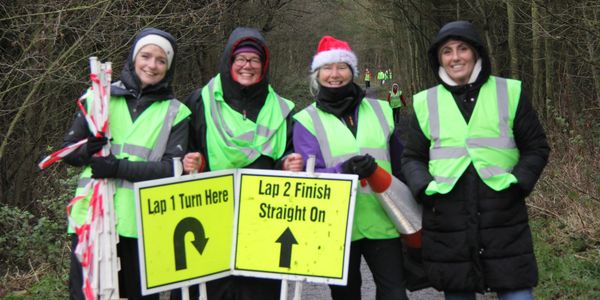 Four volunteers in reflective vests hold race direction signs in a wooded area.