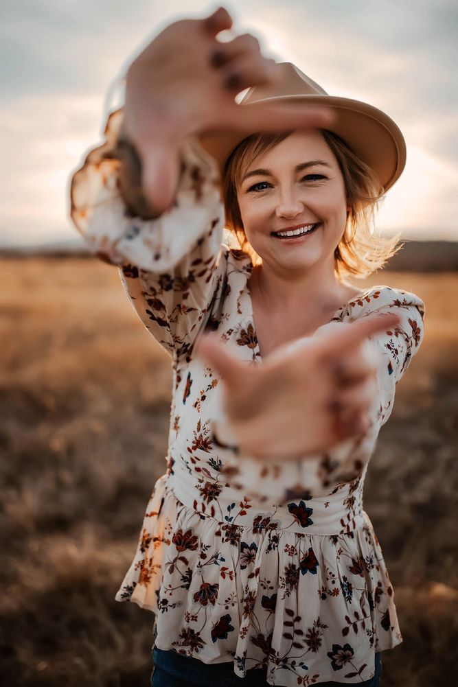 Smiling woman in a floral blouse and hat framing a shot with her hands outdoors.